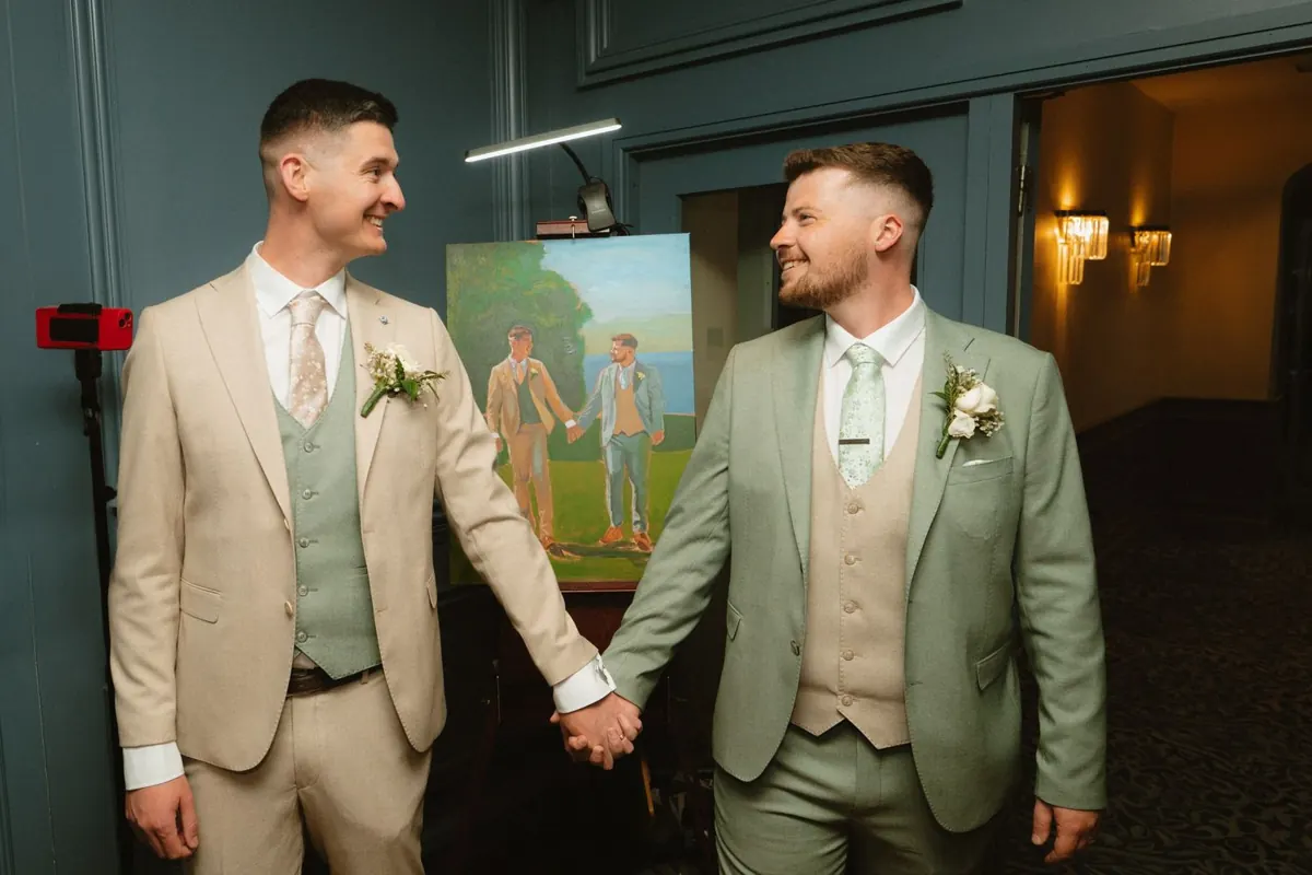 The grooms holding hands in front of a dark green doorway at night