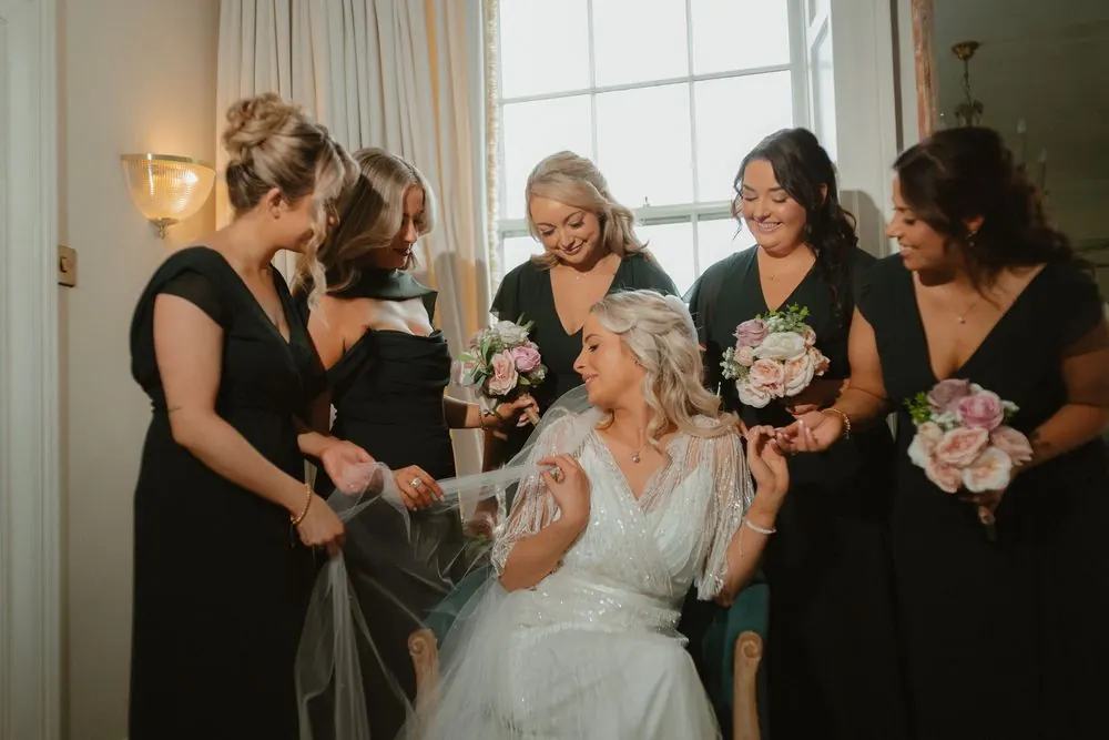 Bride in white gown sits surrounded by bridesmaids in black dresses holding pink bouquets during wedding preparations at Boyne Hill House