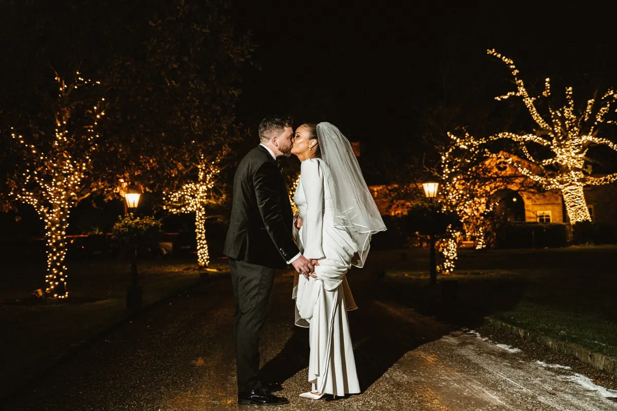 Bride and groom kissing beside illuminated trees at Clonabreany House at night.