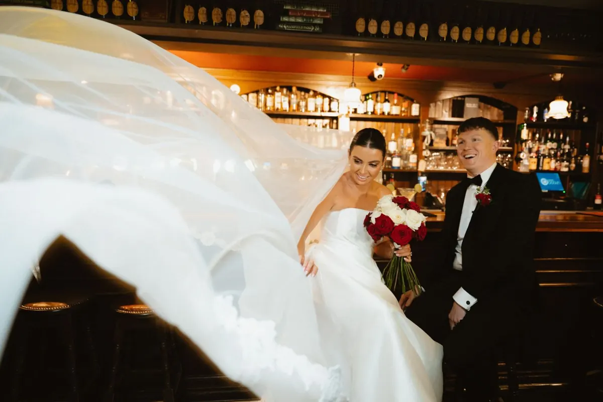 The couple standing at the wooden bar as the bride’s skirt moves across the foreground