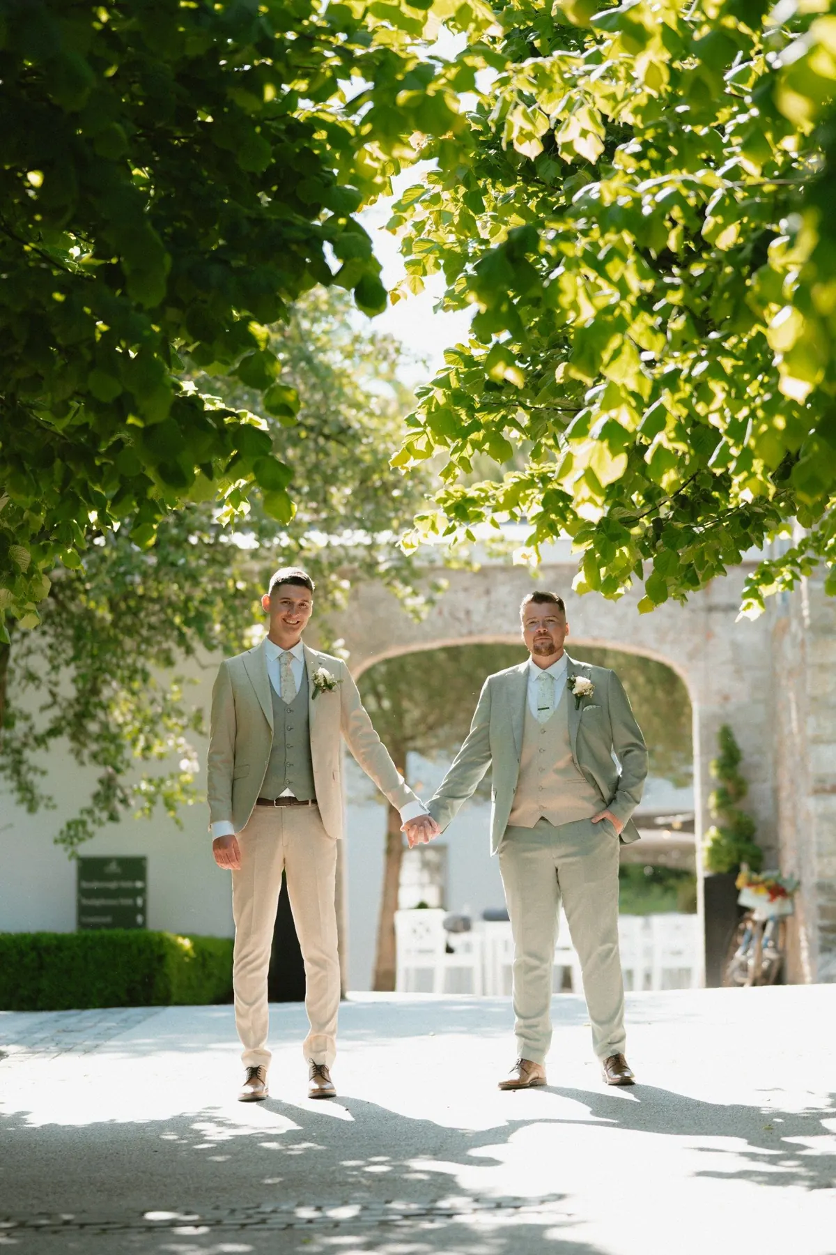 The two grooms walking hand in hand beneath leafy trees