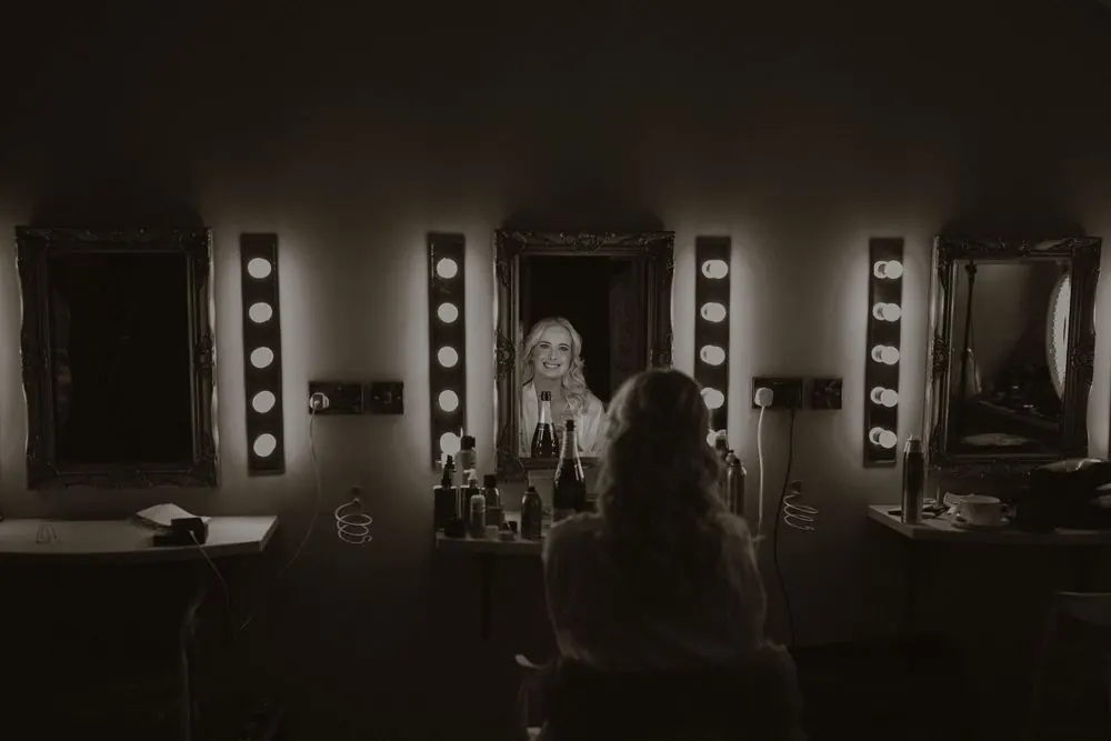 Bride sits at a vanity mirror with Hollywood lights in a dark dressing room, captured in black and white at Boyne Hill House Estate, Co. Mea