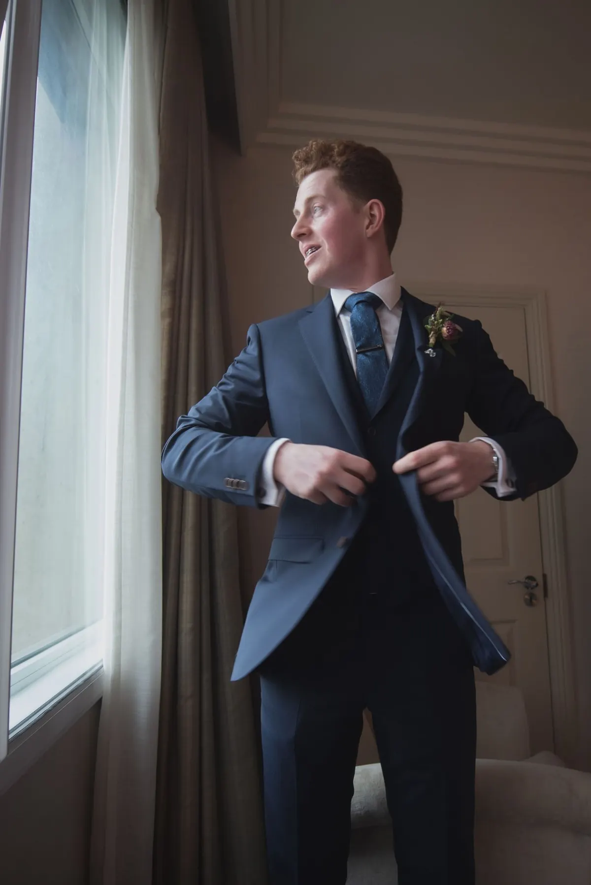 The groom adjusting his jacket beside a bright window