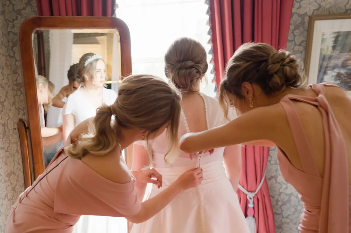 Two bridesmaids in pink dresses helping a bride with her white wedding gown in front of a mirror at Boyne Hill House, Co. Meath