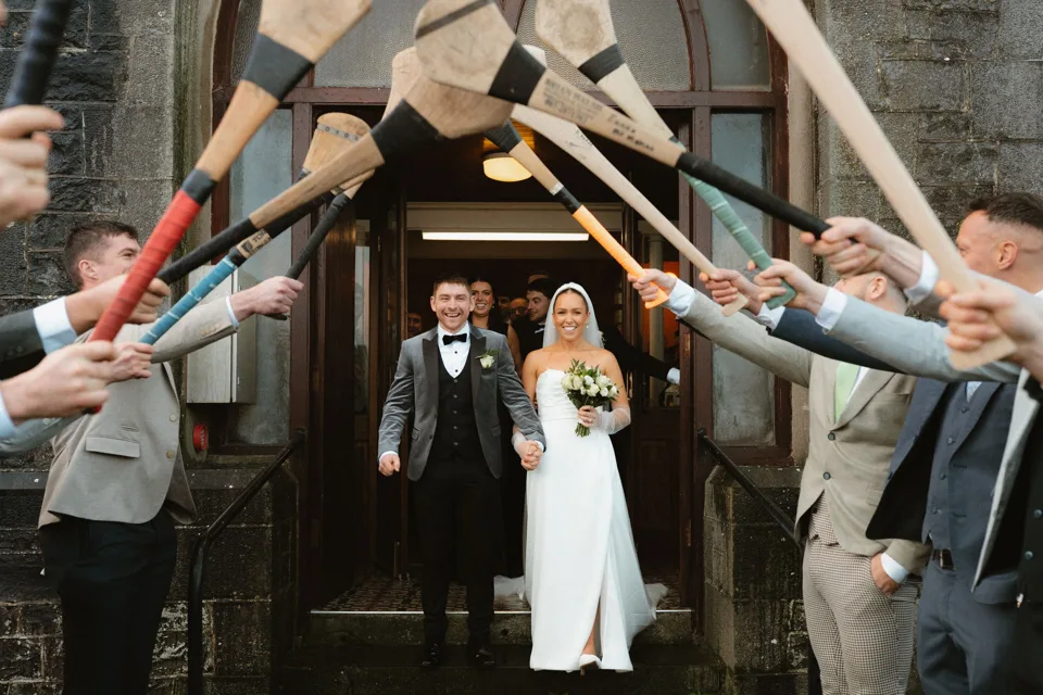 Bride and groom exit church under arch of hurling sticks held by guests at traditional Irish GAA wedding at Falls Hotel Co. Clare