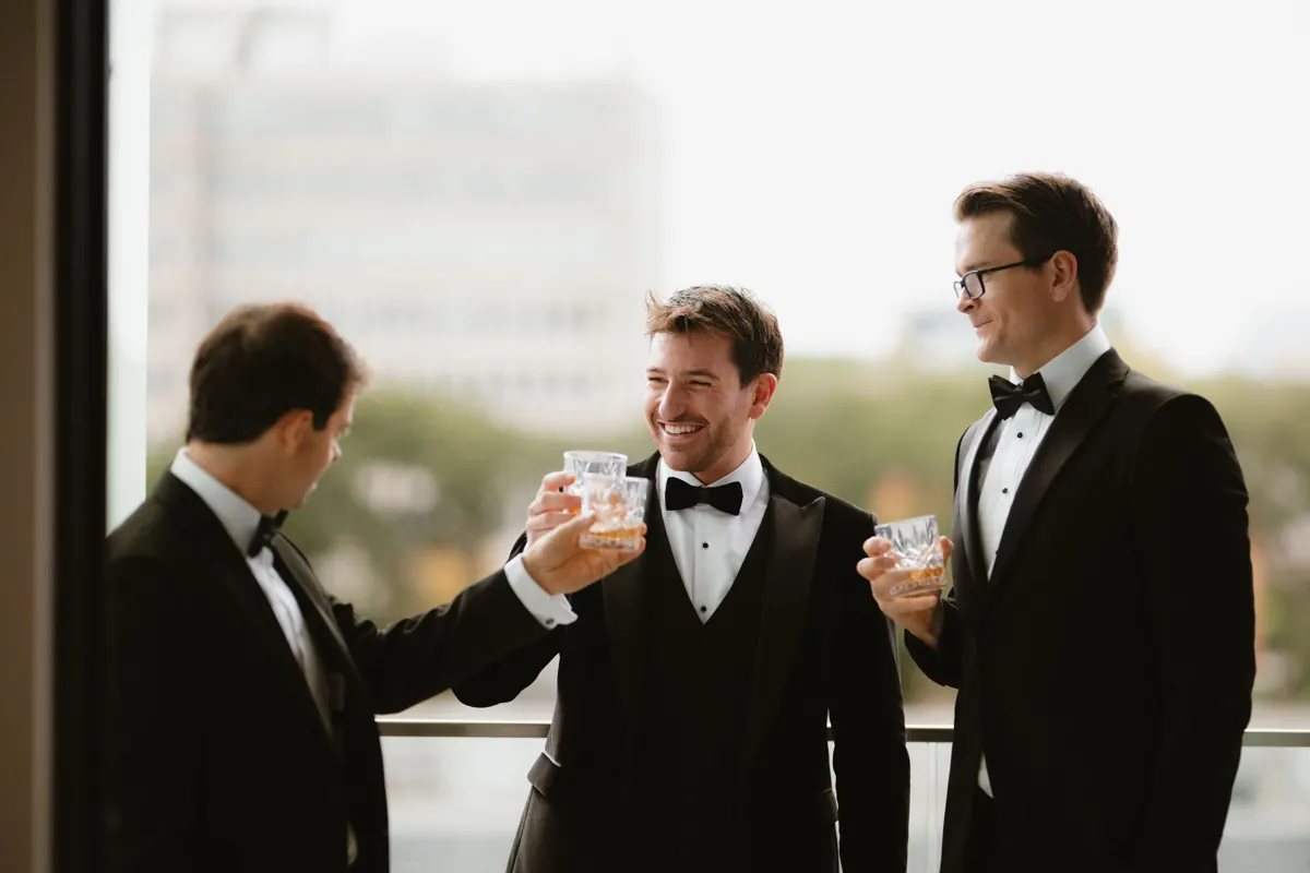 Three groomsmen in black tuxedos toasting with glasses of whiskey on a balcony during a wedding celebration at the Grayson Co. Dublin
