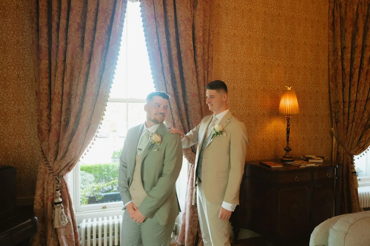 The grooms standing beside a tall window and red curtains