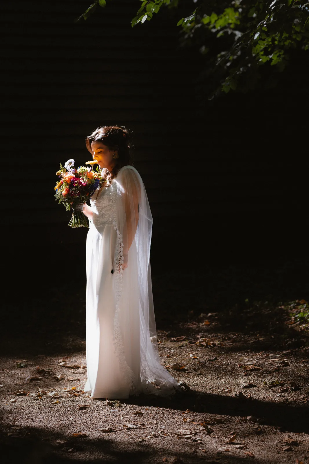 Aoife holds her bouquet in a patch of light beside the dark timber barn wall.