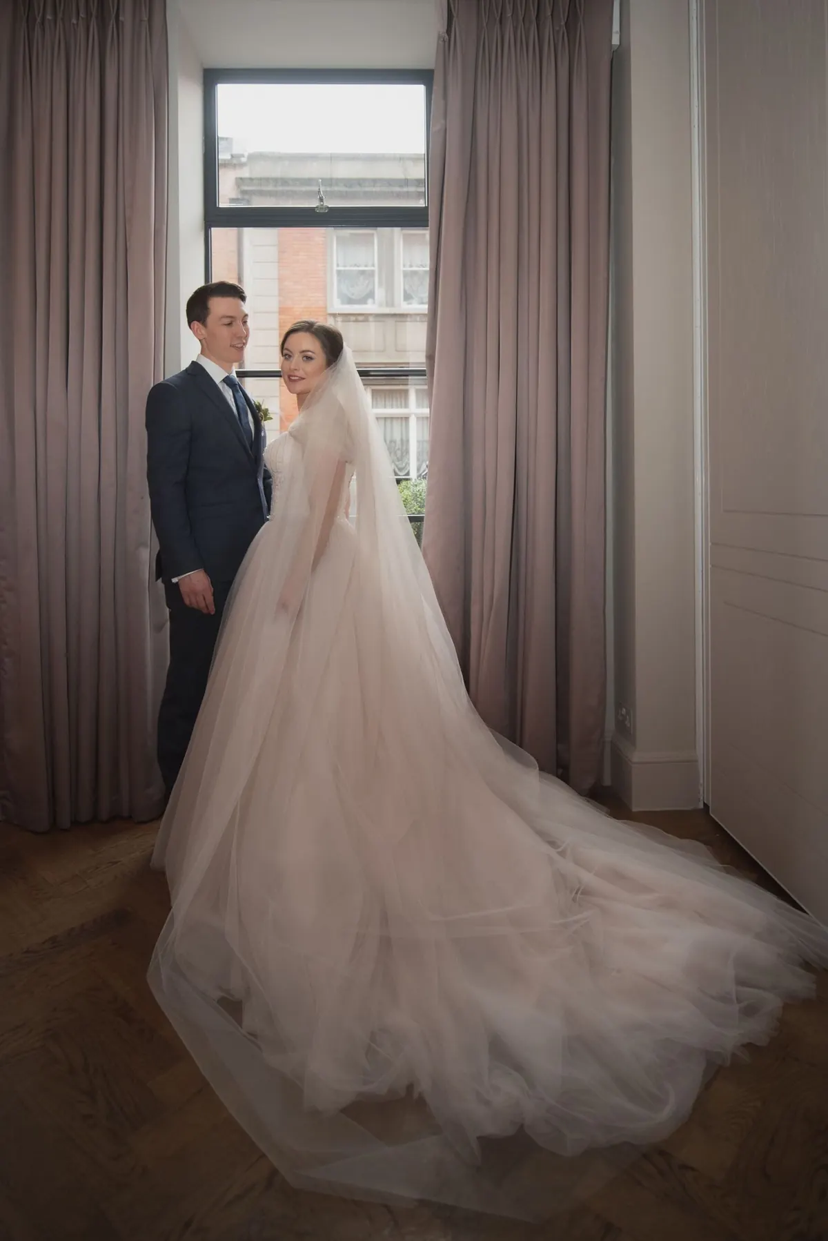 The couple standing together beside a tall window