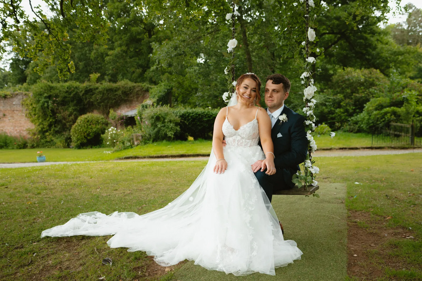 Claire sits on a flower decorated garden swing beside Will, with her dress spread across the grass.
