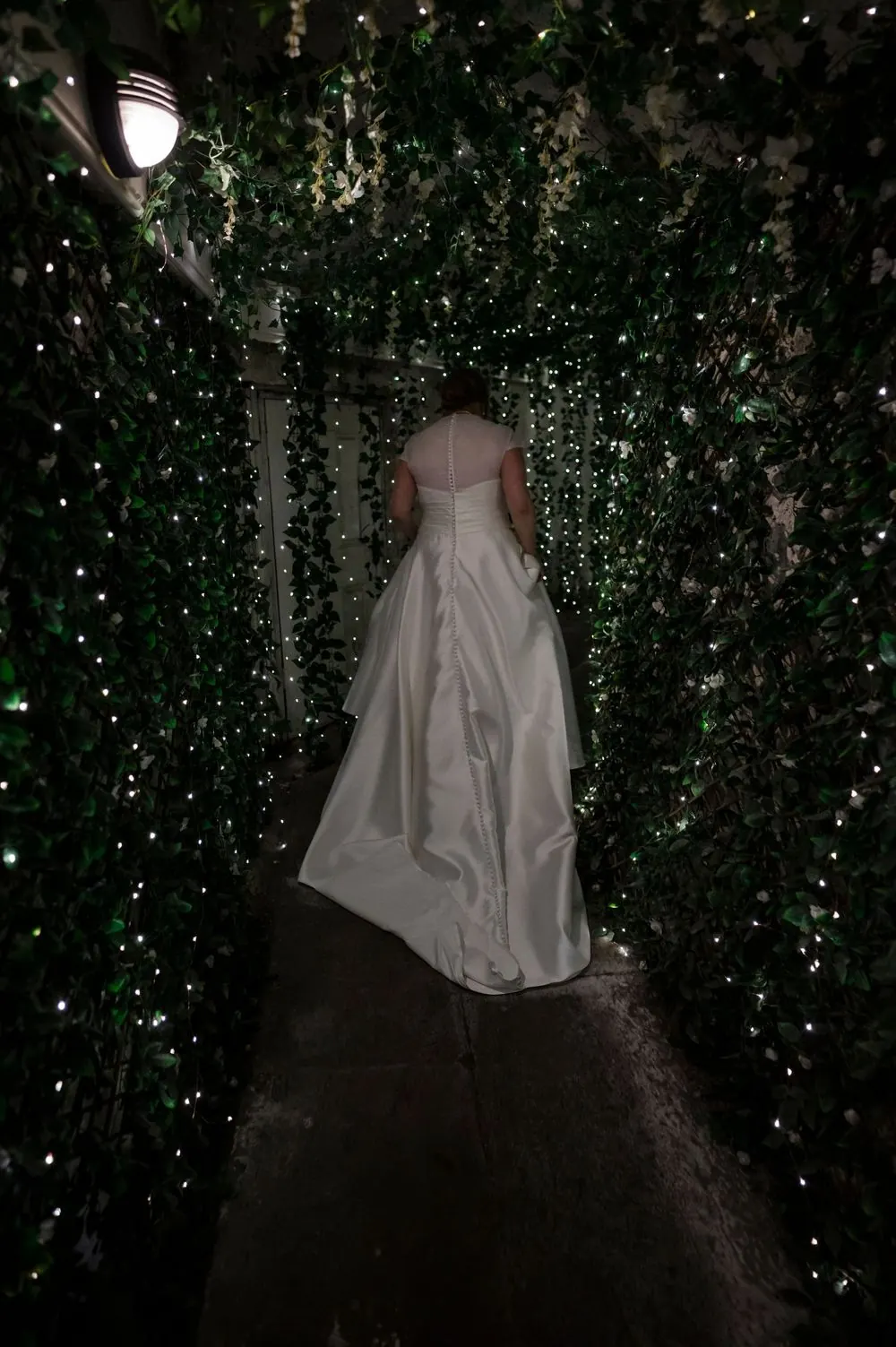 Bride in white wedding gown walking through a magical ivy tunnel adorned with twinkling fairy lights at Boyne Hill House Estate, Co.Meath