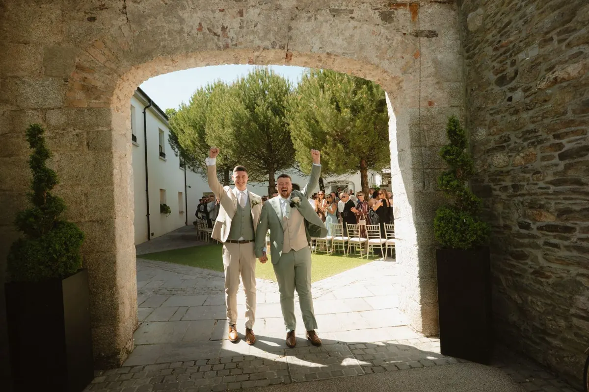 The grooms framed by the stone arch beside the outdoor ceremony