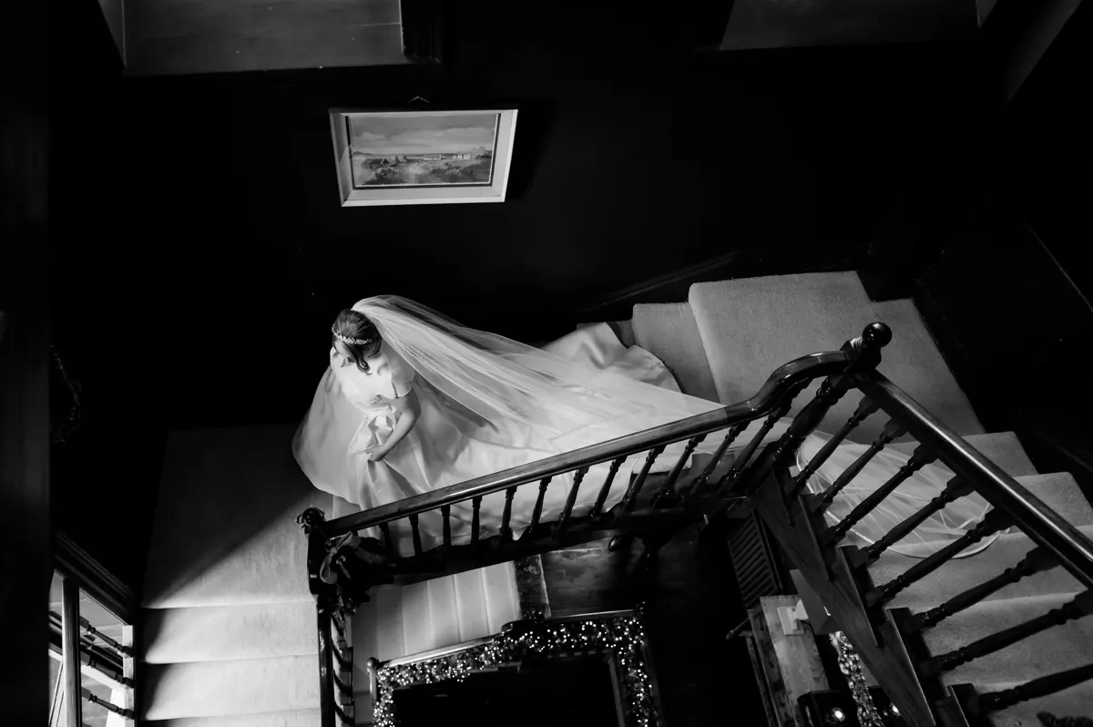Black and white photo of a bride in a flowing gown and veil descending an elegant wooden staircase at Boyne Hill House, Co. Meath