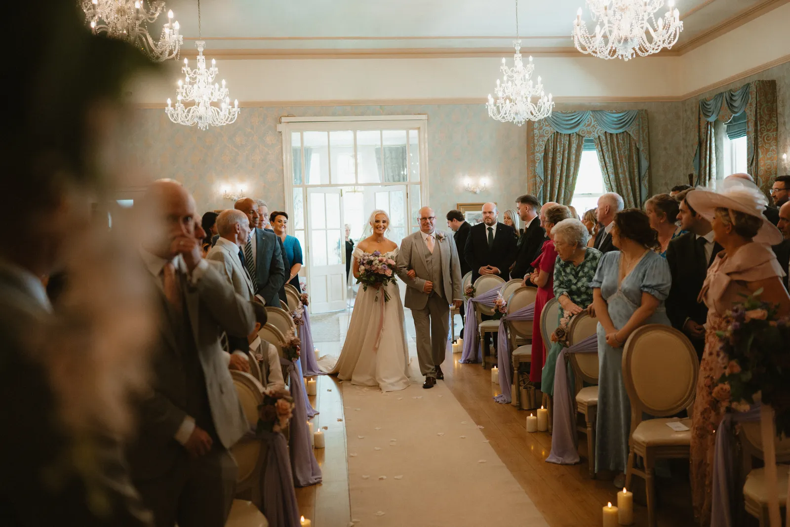 Emma enters Corick House’s chandelier lit ceremony room between rows of standing guests.