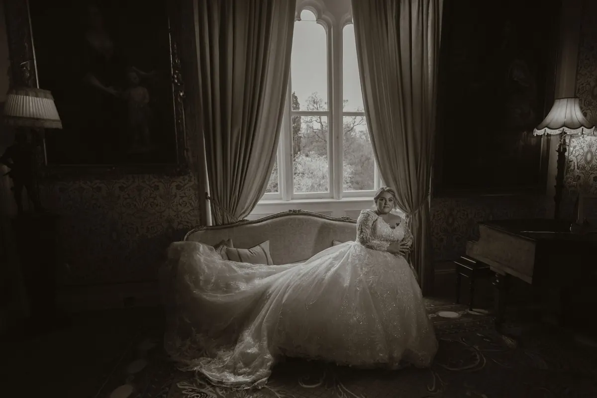 Bride in a white ball gown sits gracefully on a vintage sofa by a grand window in a dimly lit elegant room at Cabra Castle