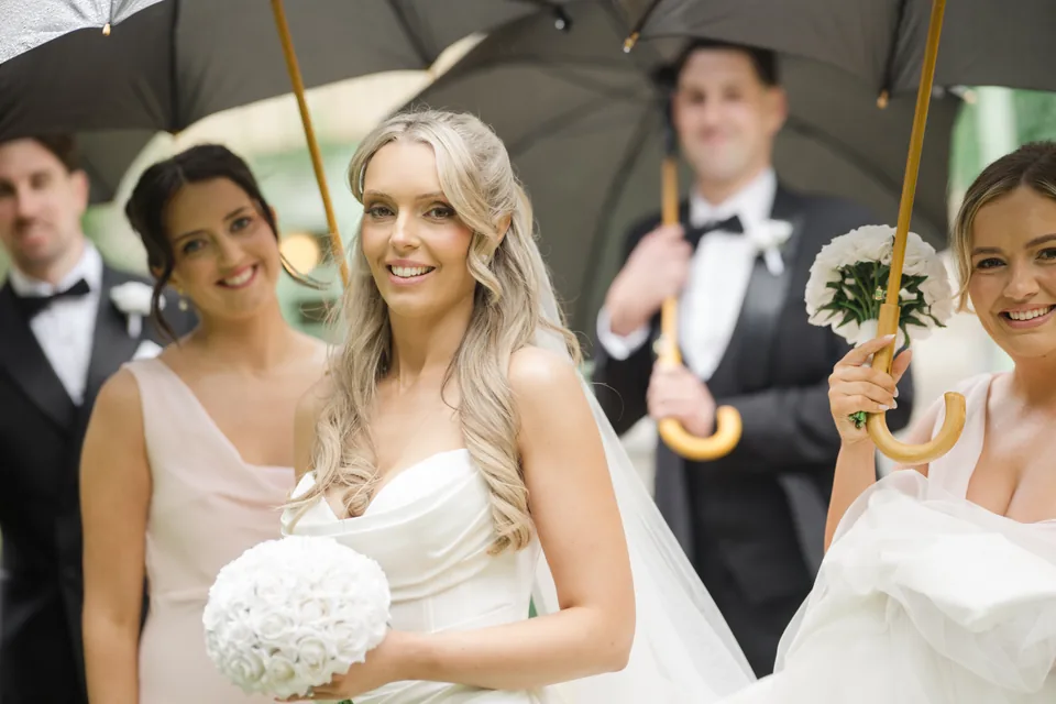 The bridal party laughs together beneath the umbrellas.