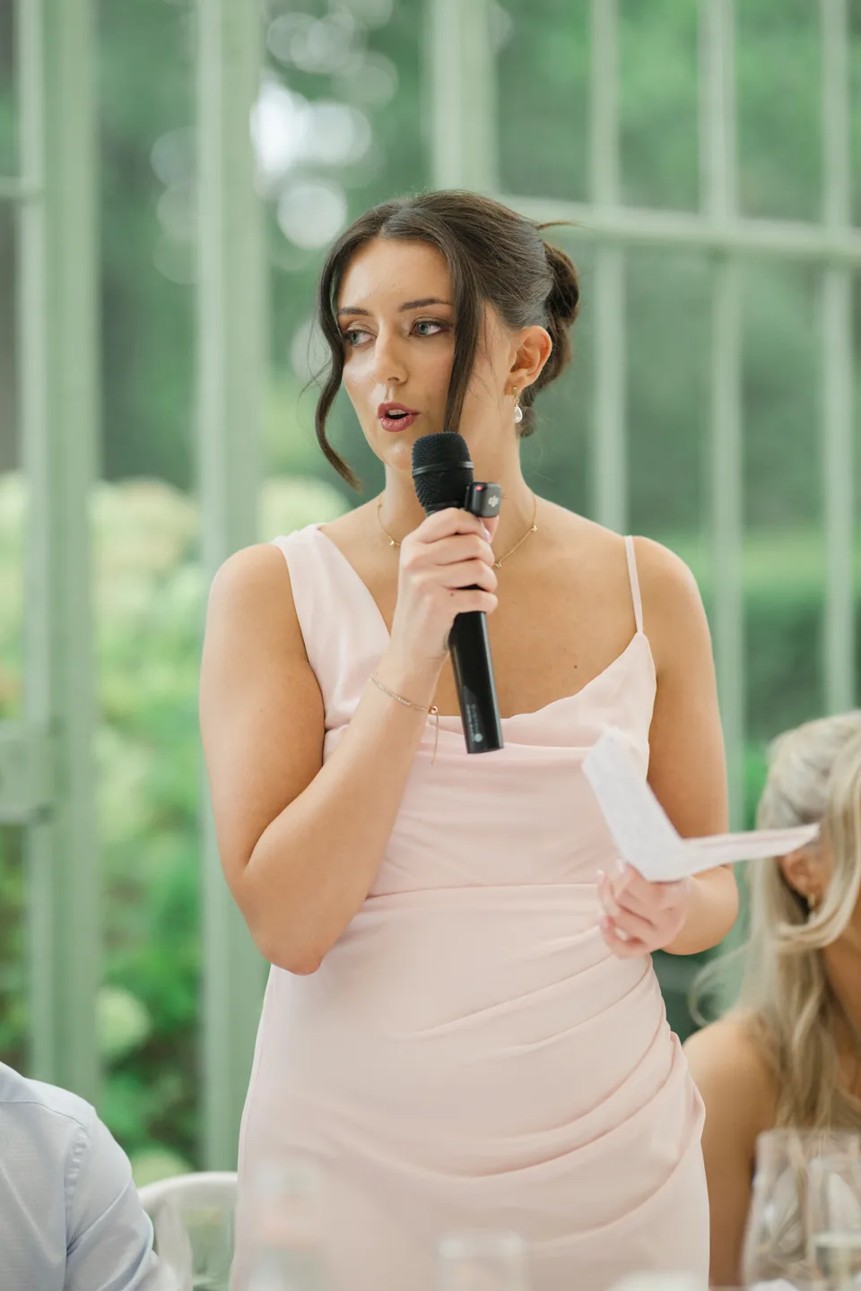A wedding speech beside the long table in the glass-roofed room.