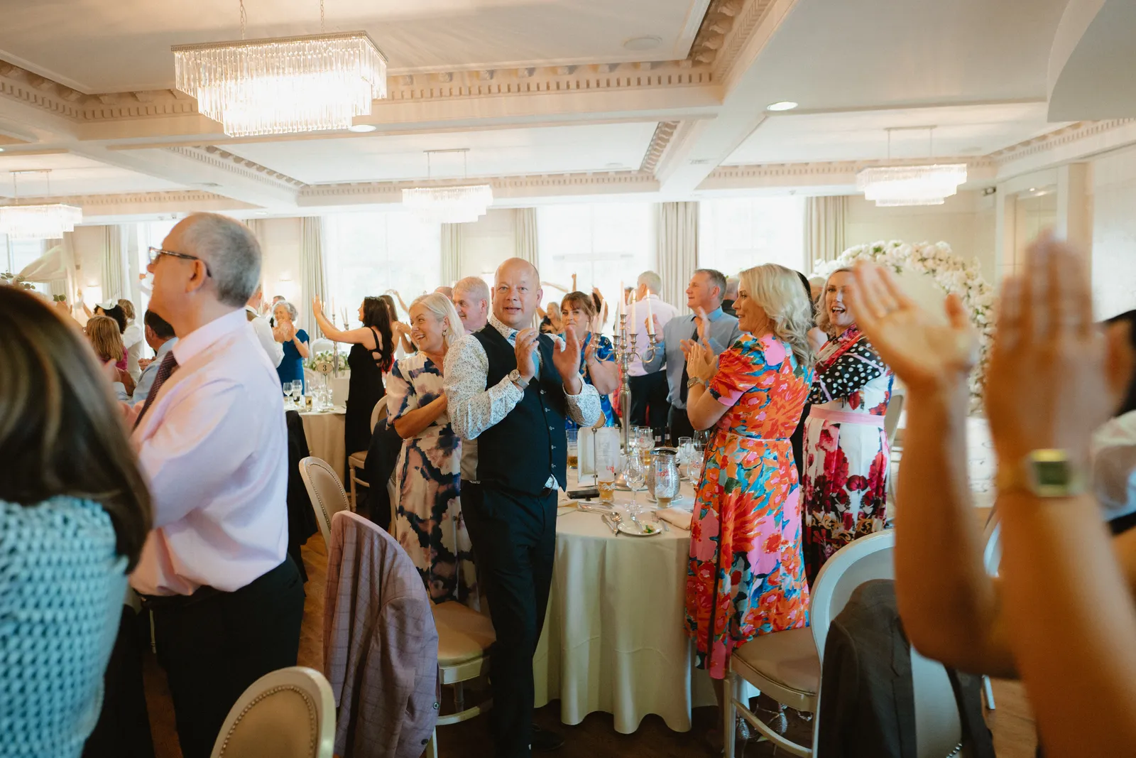 Guests stand and applaud around the reception tables beneath rectangular chandeliers.