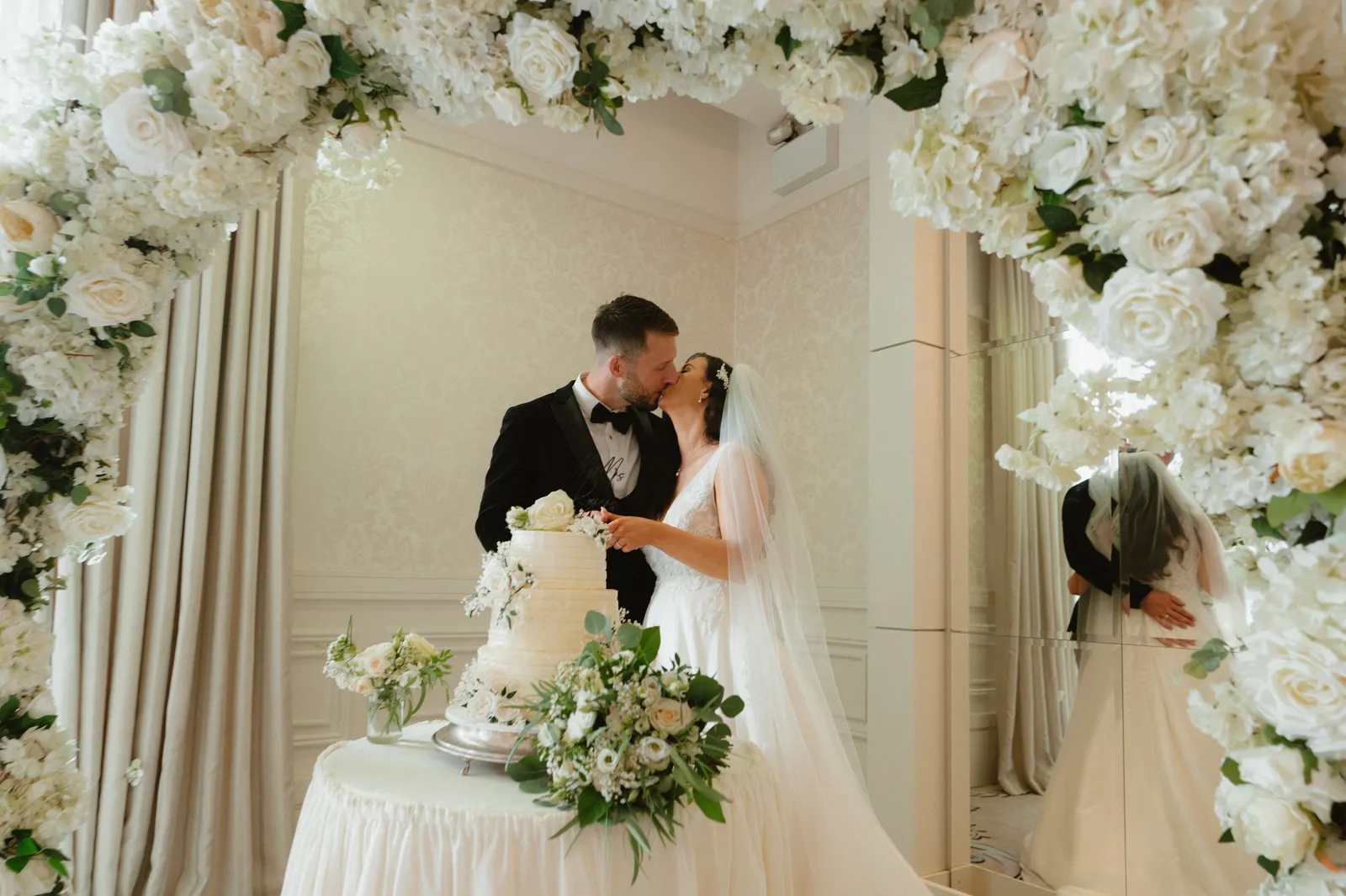 Kerstin and Cian kiss beside their tiered cake beneath a white flower arch.