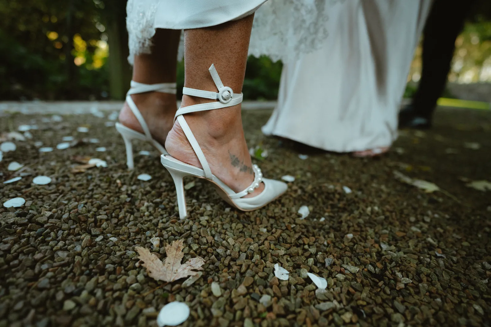 Nicola’s white strappy heels and dress hem on a gravel path scattered with white petals.