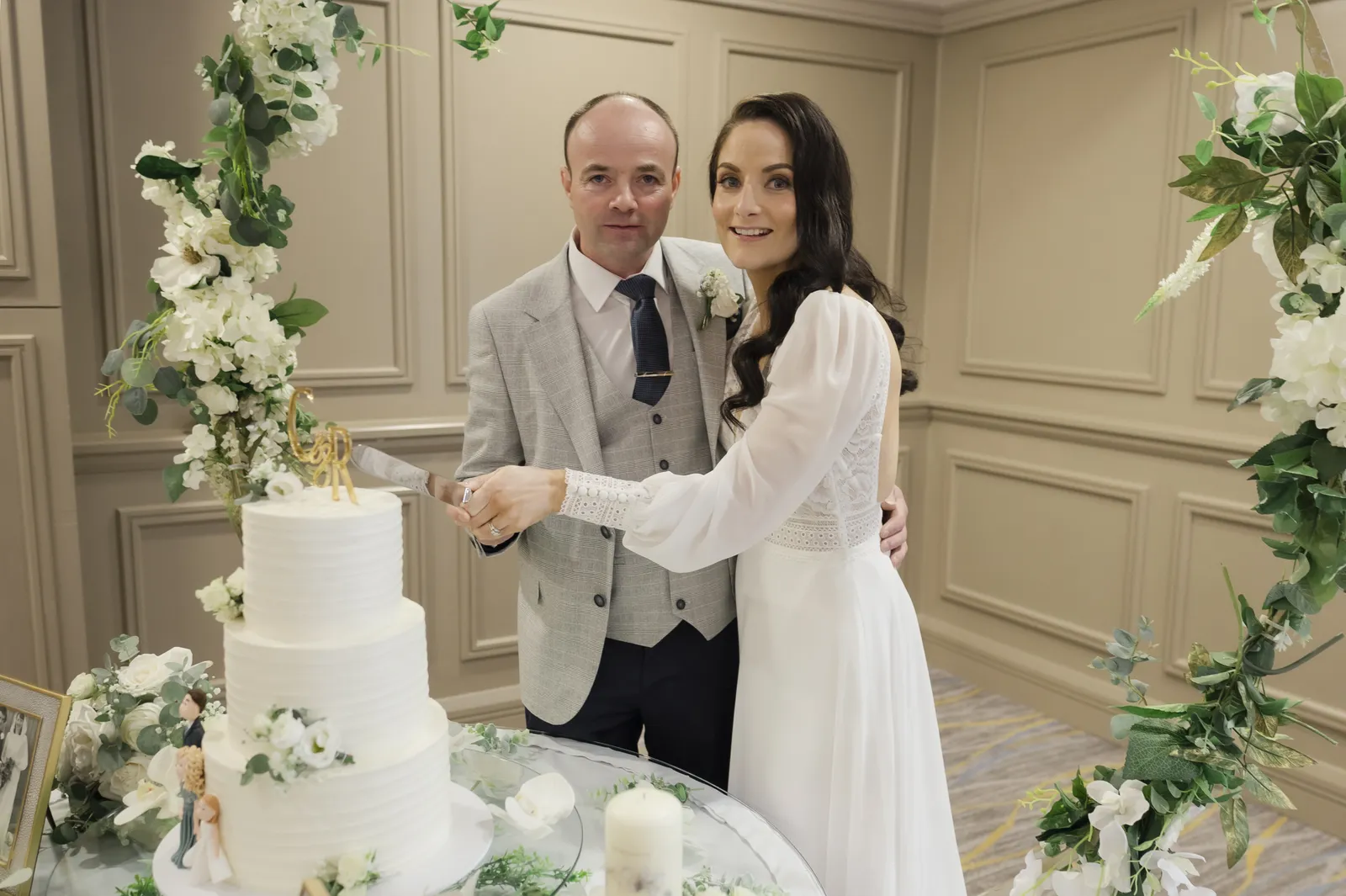 Roisin and Colm cut their white tiered cake between tall arrangements of flowers and greenery.
