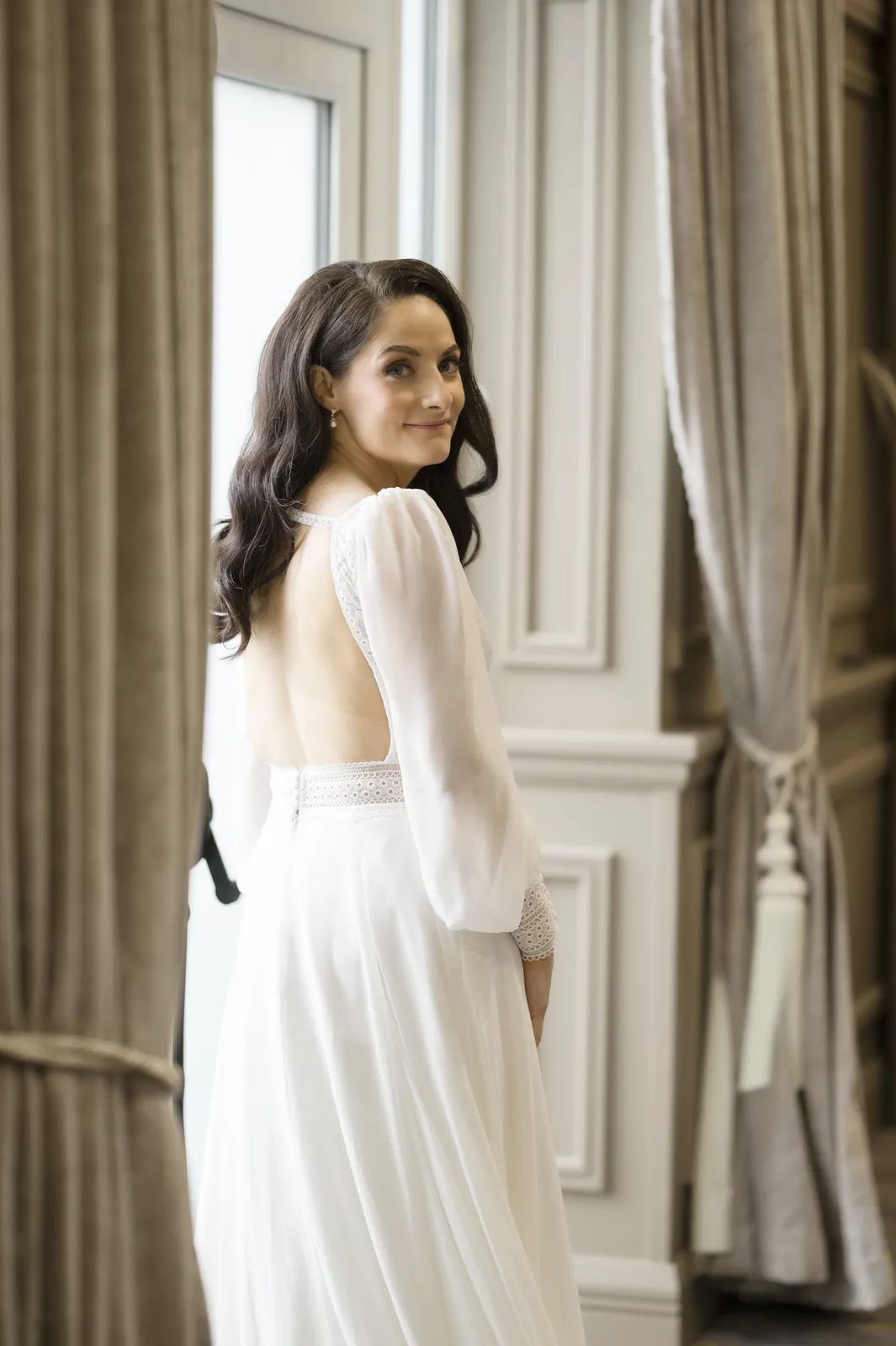 Roisin looks over her shoulder beside a tall window framed by heavy curtains.