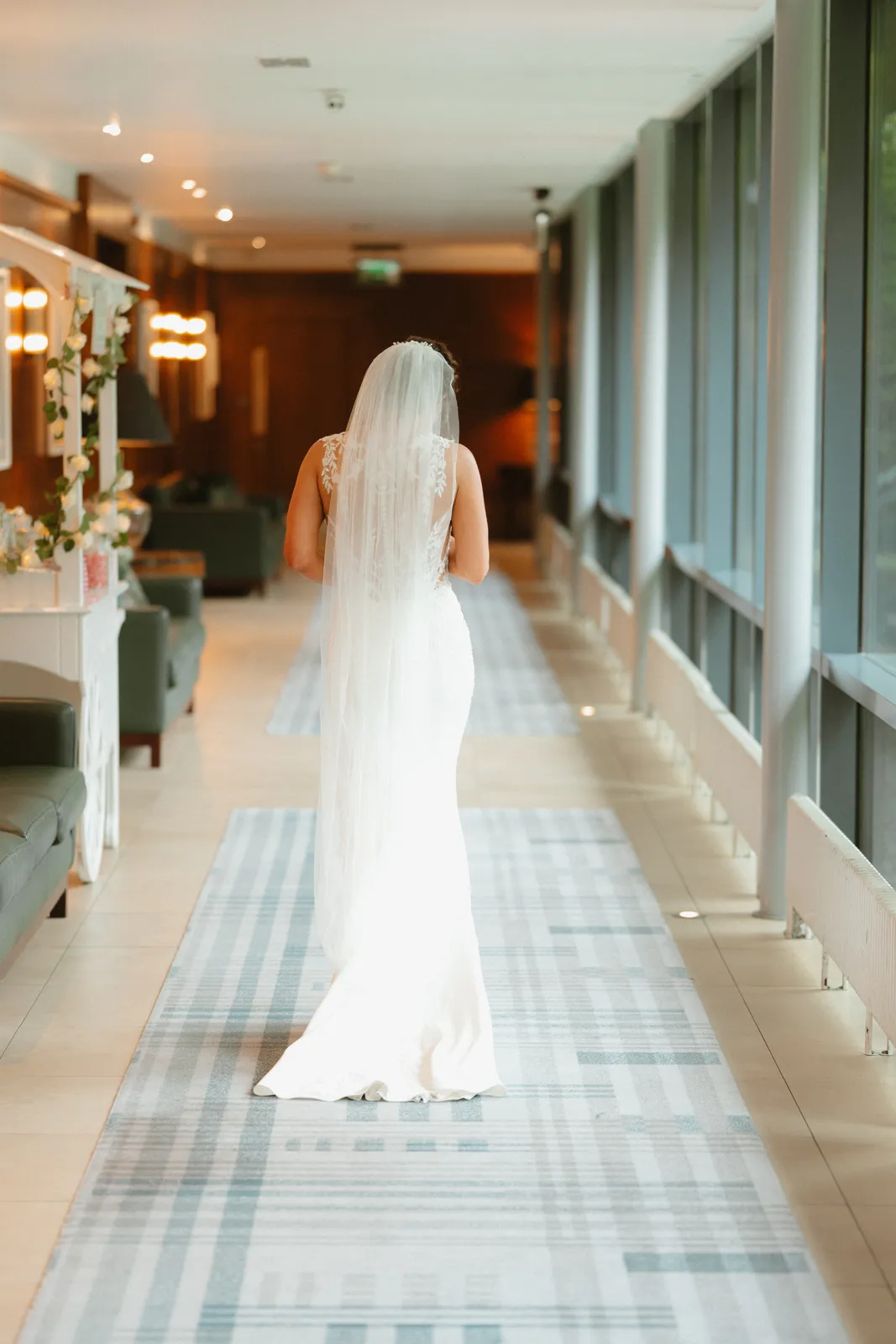 Vicky walks along a bright window lined corridor at Ballymascanlon, her veil and dress train behind her.