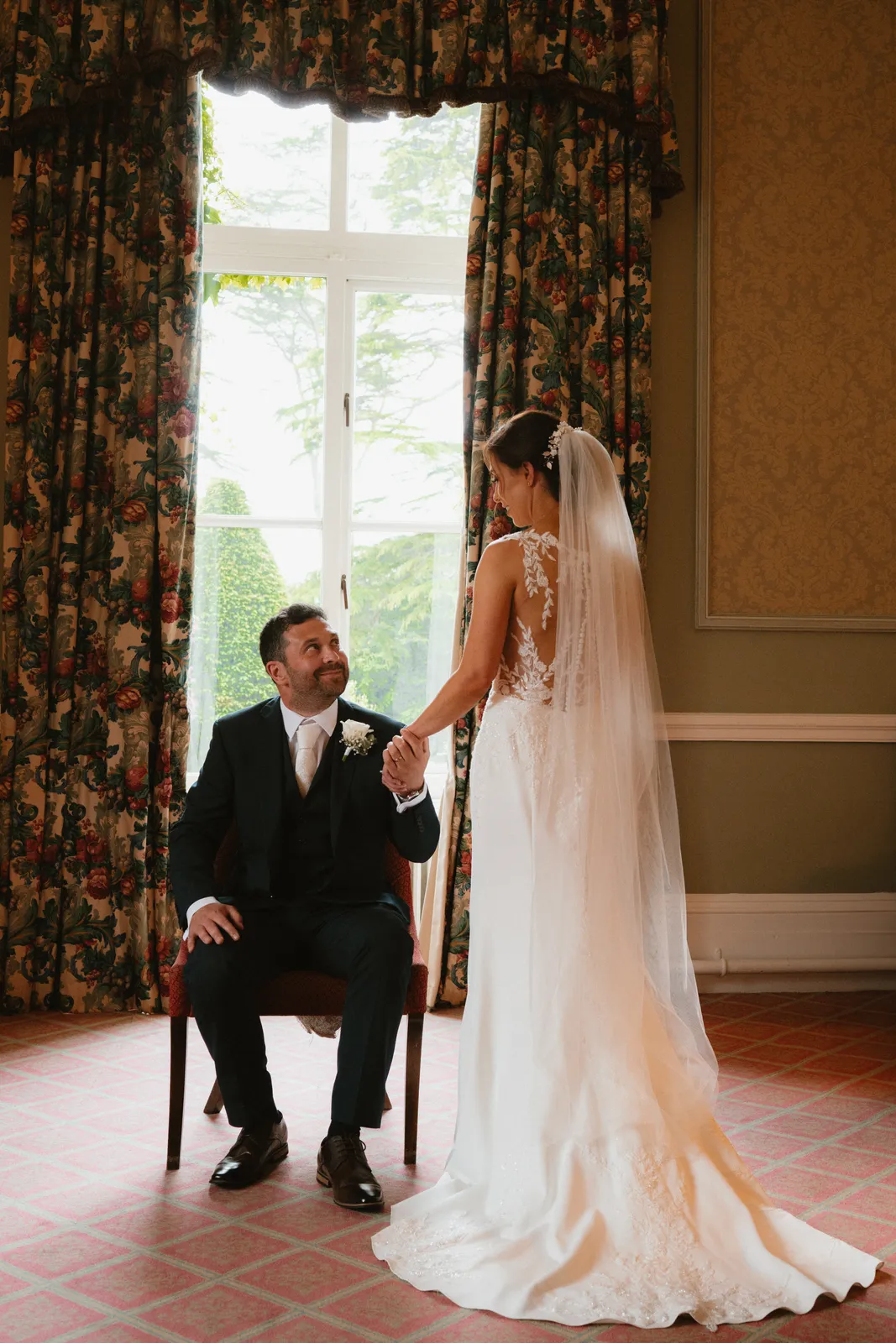Pairic sits beside a tall curtained window and holds Vicky’s hand as she stands beside him.